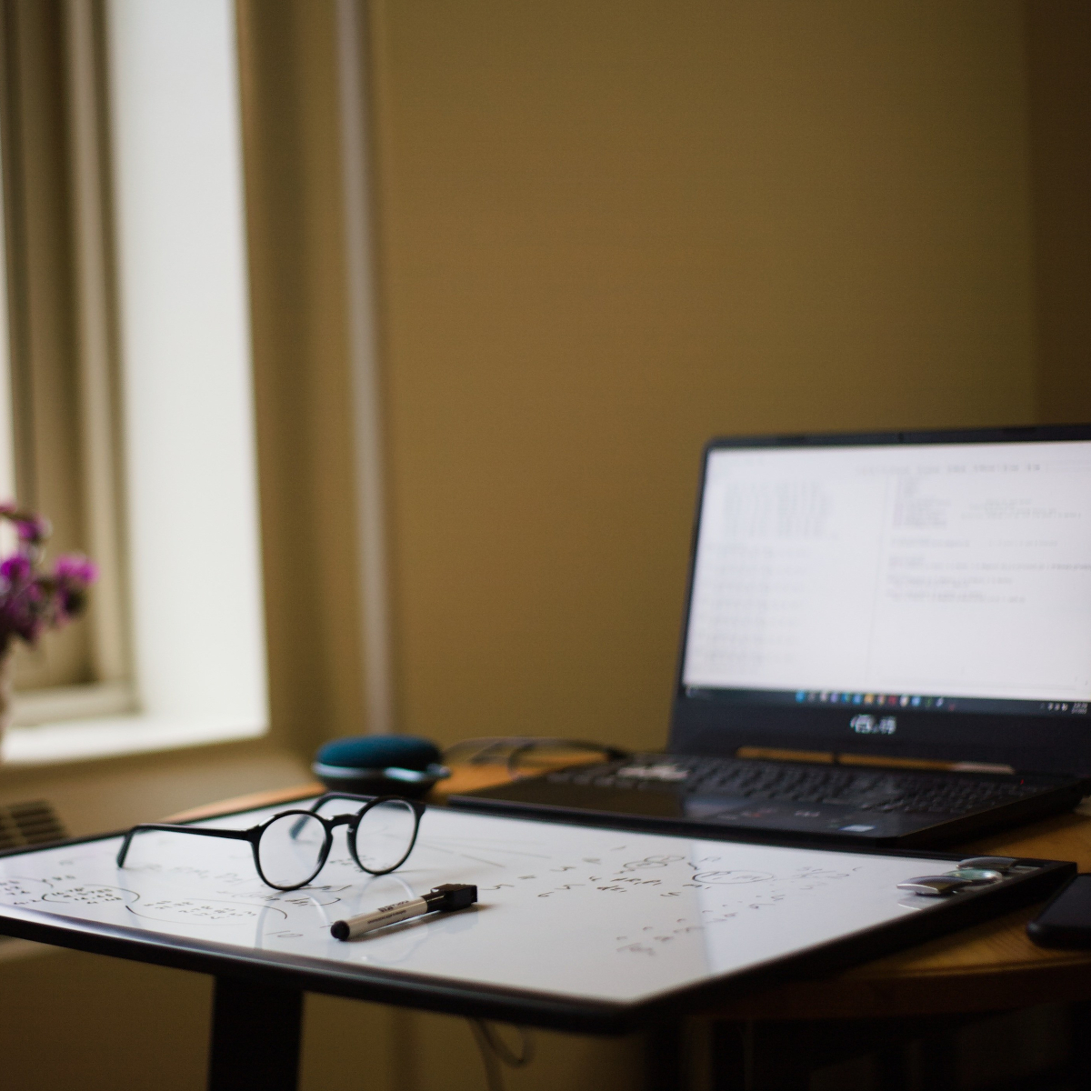 Na zdjęciu widać biurko w gabinecie, na którym leżą okulary, marker i tablica lub arkusz z odręcznymi notatkami. W tle znajduje się otwarty laptop, a wnętrze ma ciepłe, stonowane oświetlenie.